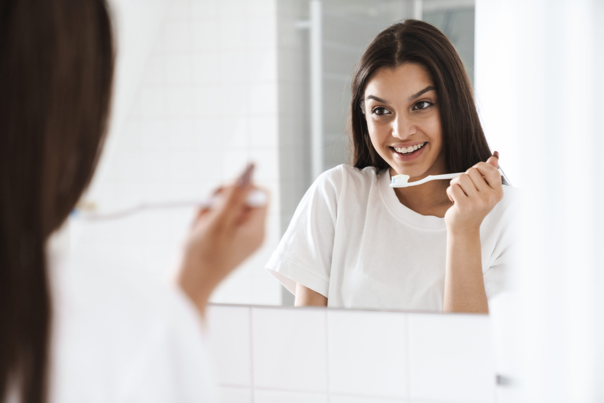 Woman brushing her teeth in the mirror following a consistent daily oral care routine